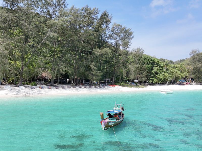 A moored longtail boat over bright turquoise water in front of a tree-lined beach lined with sun loungers