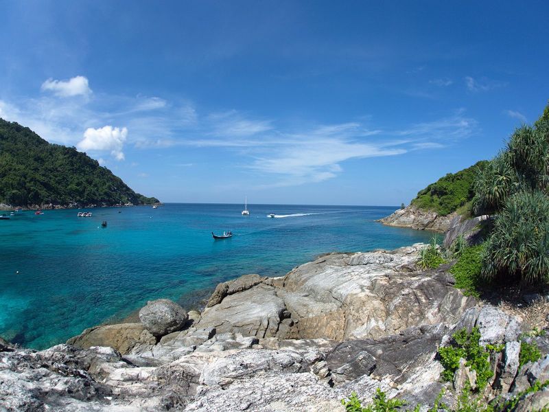 Rocky shoreline framing a calm blue bay dotted with longtail boats and a catamaran at Racha Yai