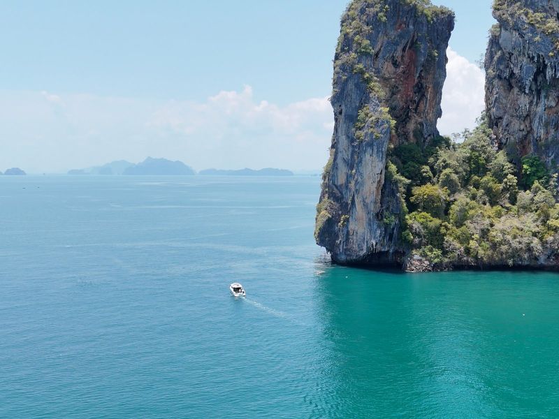 A speedboat passing beneath a sheer limestone cliff on the open water of Phang Nga Bay